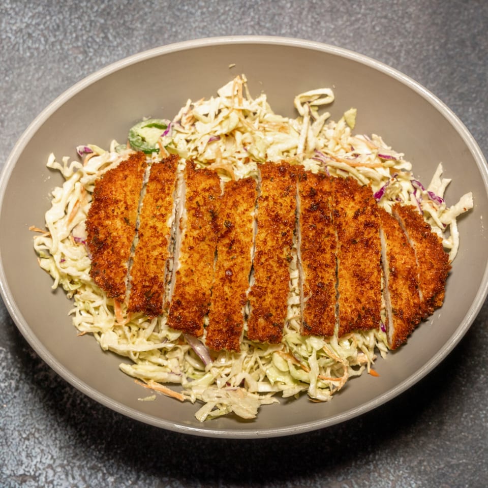 Crispy fried chicken cutlet, sliced and served over a bed of creamy coleslaw in a gray bowl.