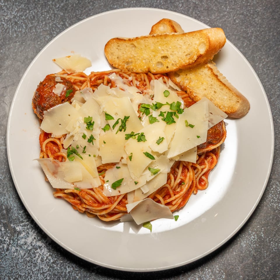 Spaghetti and meatballs with tomato sauce, topped with shaved parmesan and parsley, served with garlic bread.