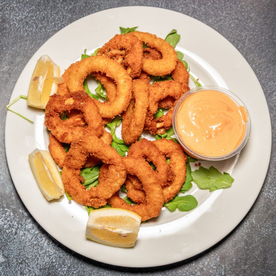 Crispy fried calamari rings with lemon wedges and a side of dipping sauce on a white plate.