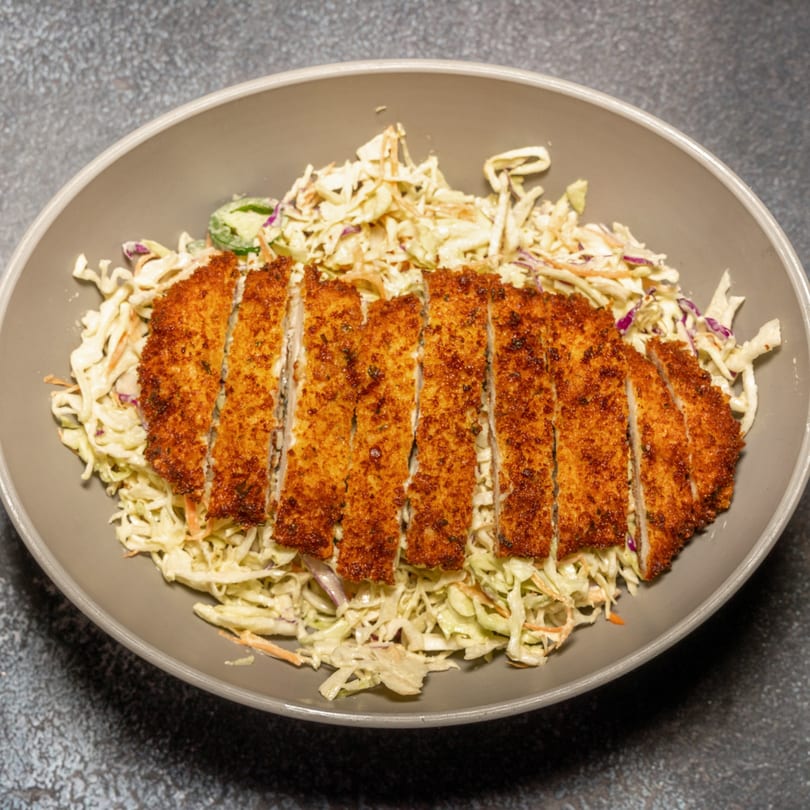 Crispy fried chicken cutlet, sliced and served over a bed of creamy coleslaw in a gray bowl.